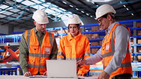 Two men and a woman in high visibility vests and hard-hats examining a laptop in a warehouse.